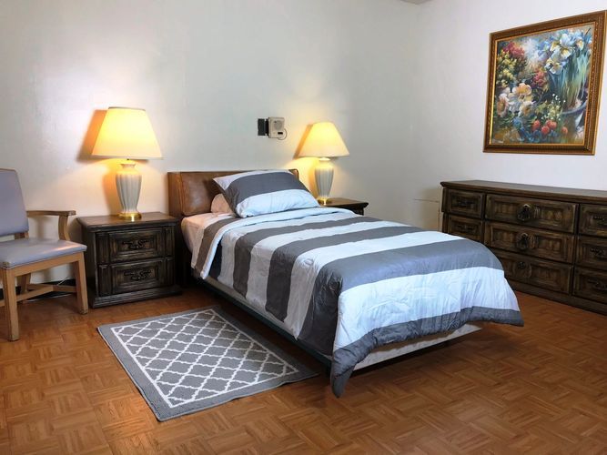 A cozy bedroom with a single bed, flanked by two nightstands and lamps, featuring a patterned rug and a dresser.