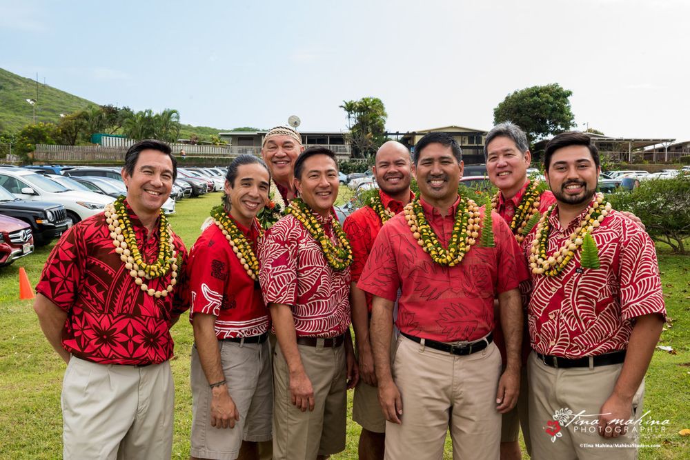 A group of men wearing red shirts and leis, smiling outdoors in Hawaii.