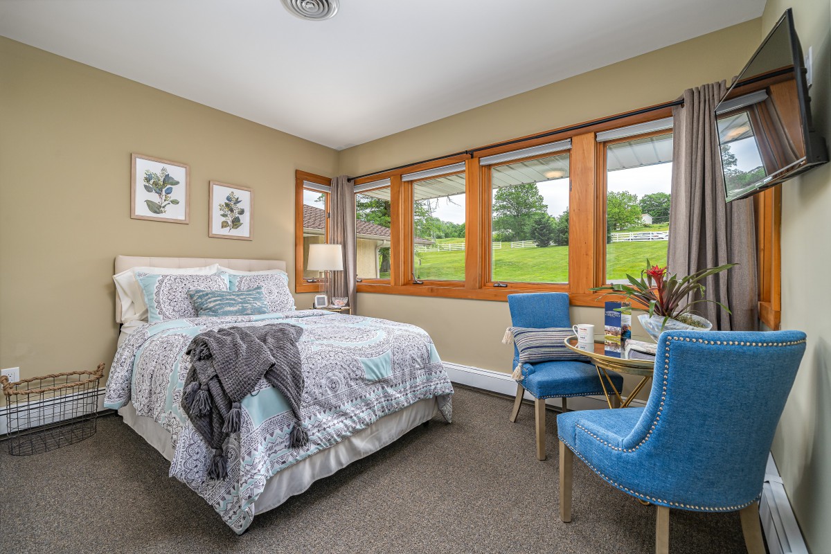 A cozy bedroom with a large window overlooking a green landscape, featuring a bed and blue chairs.