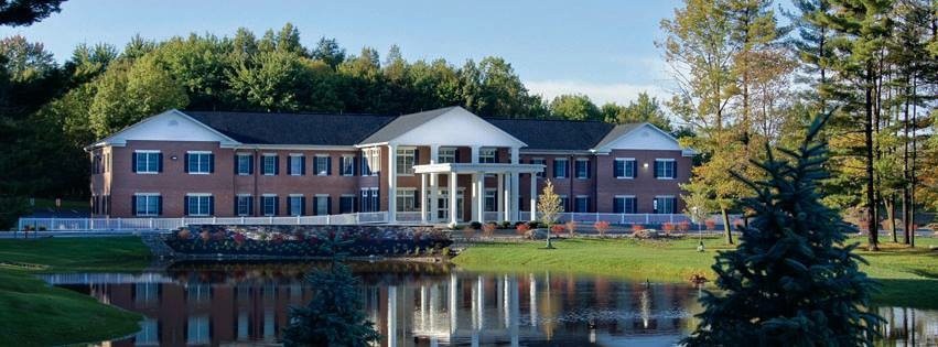 A beautiful building with a white facade and large windows is reflected in a calm lake, surrounded by lush greenery.