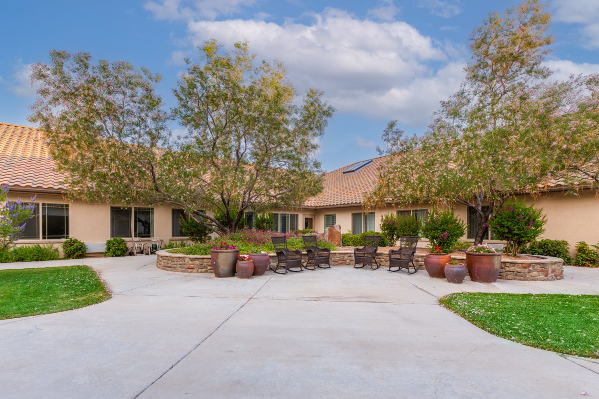 A peaceful courtyard with outdoor seating, surrounded by trees and potted plants under a partly cloudy sky.