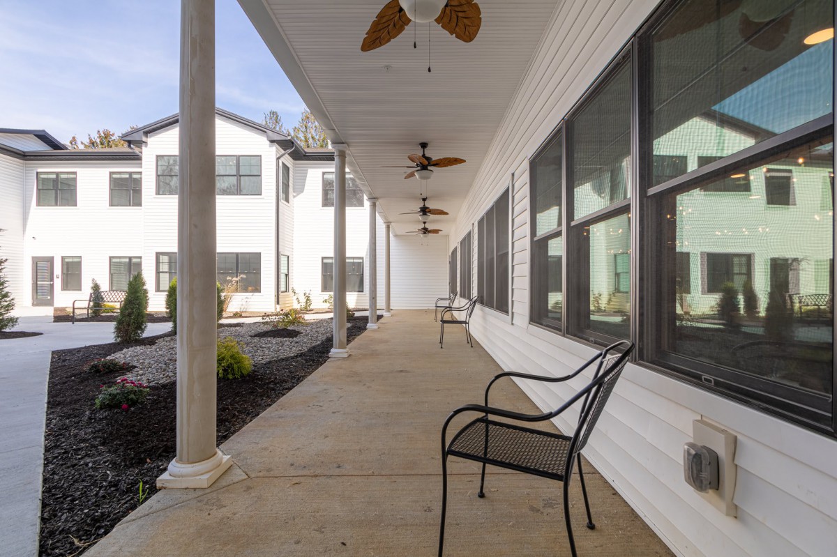 A long porch with chairs, ceiling fans, and large windows along a white building.