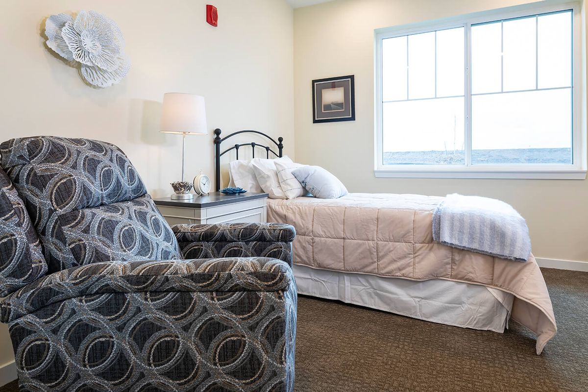 A cozy bedroom featuring a bed with white linens, an armchair, a lamp on a side table, and a large window.