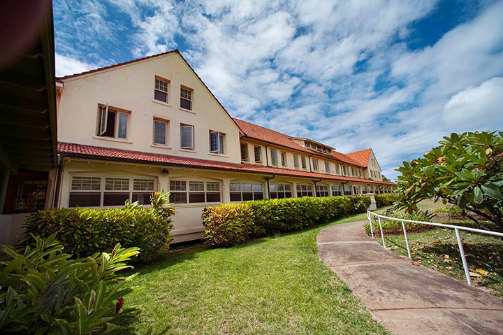 A scenic view of a long building with a red roof, surrounded by greenery under a partly cloudy sky.