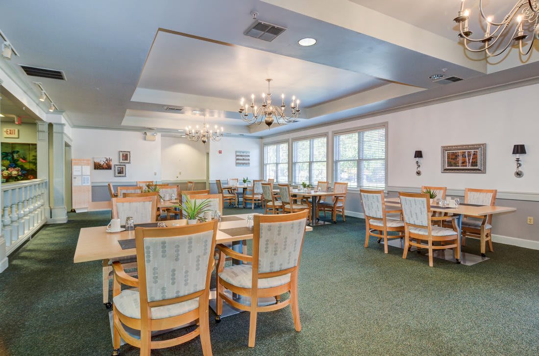 A well-lit dining room with multiple tables and chairs, featuring a chandelier and large windows.