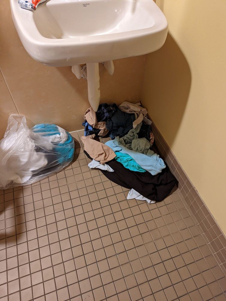 A cluttered bathroom corner with clothes on the floor beneath a sink.