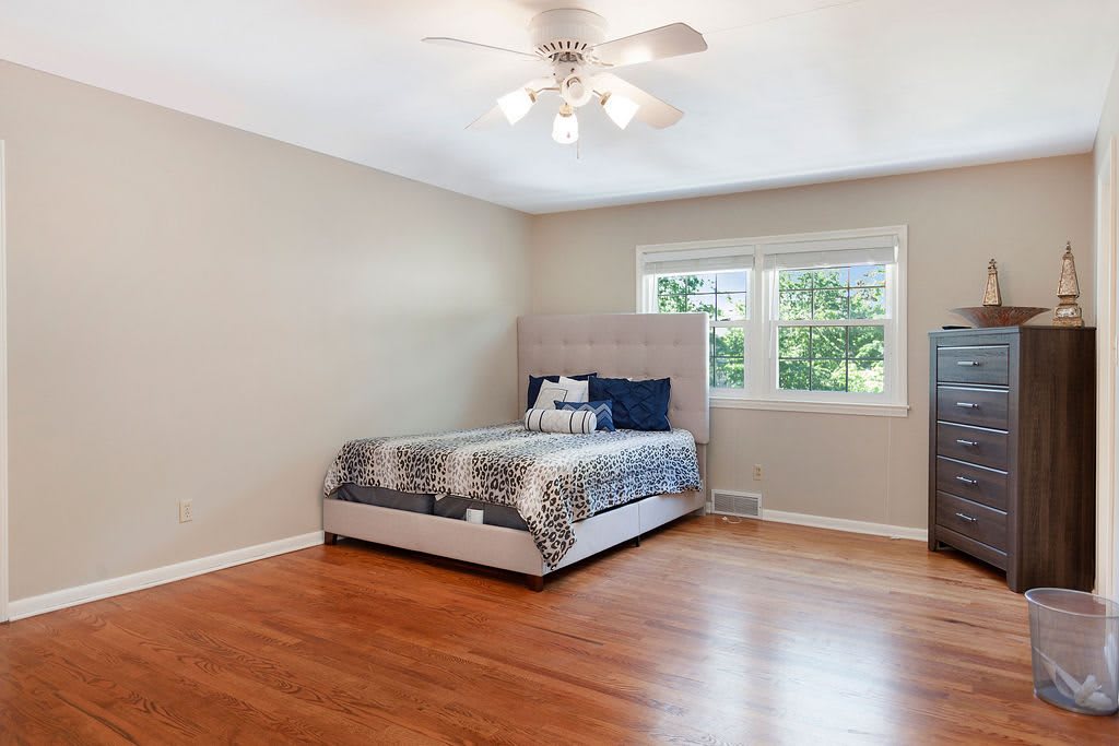 A spacious bedroom with a simple bed, hardwood floors, and a ceiling fan.