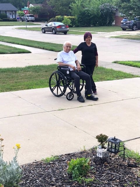 A caregiver stands beside an elderly person seated in a wheelchair on a residential sidewalk.
