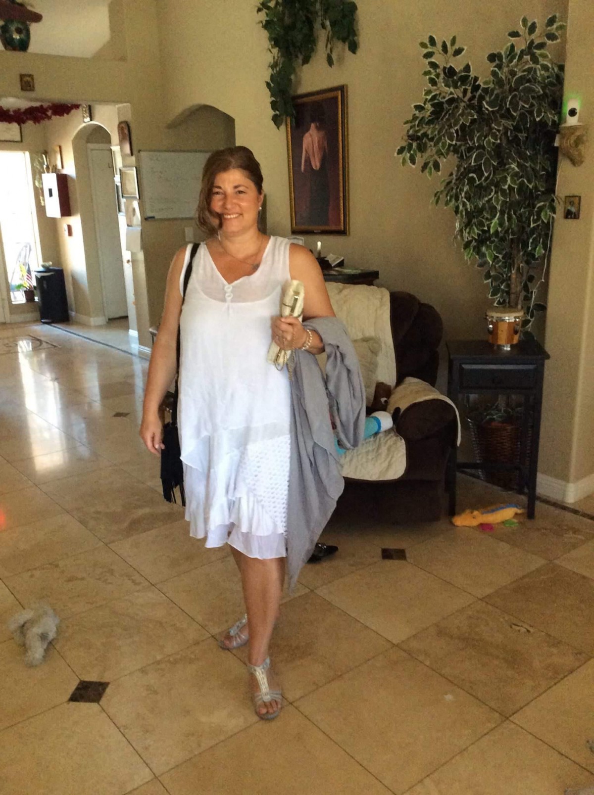 A woman in a white dress stands in a living room holding some items.