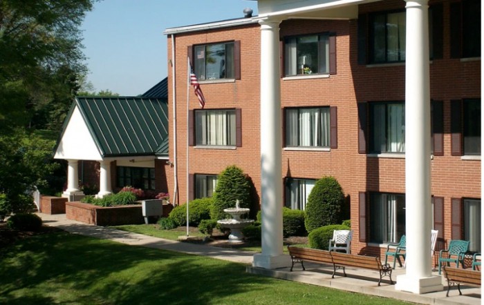 A brick building with large windows, flanked by columns and surrounded by well-maintained greenery and benches.