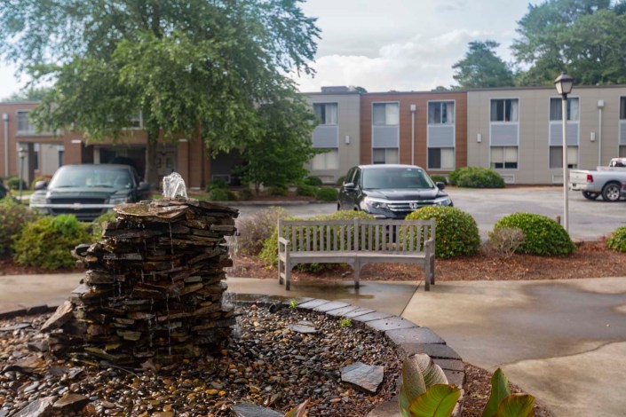 A small fountain and bench situated in front of an apartment complex parking lot.