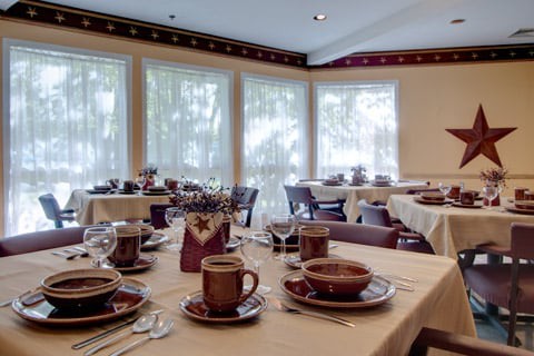 A cozy dining room with neatly set tables, featuring brown cups and plates near large windows.
