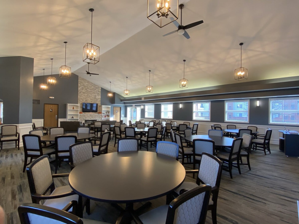 A spacious dining area with multiple tables and chairs, elegantly lit by modern chandeliers, featuring large windows and ceiling fans.