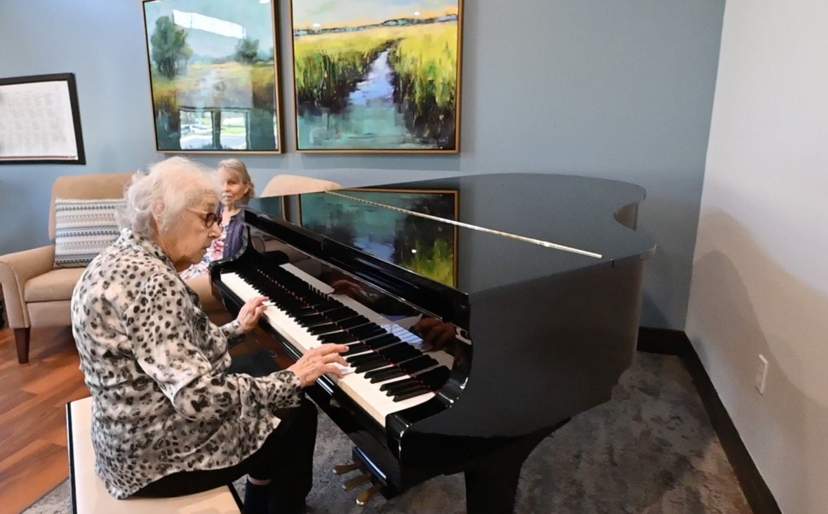 An elderly woman playing the piano in a room with artwork on the walls.