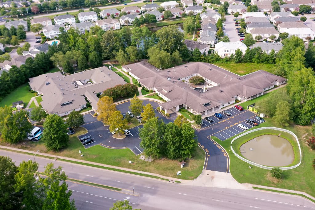 Aerial view of a building complex surrounded by residential homes and greenery, featuring a parking lot and nearby road.