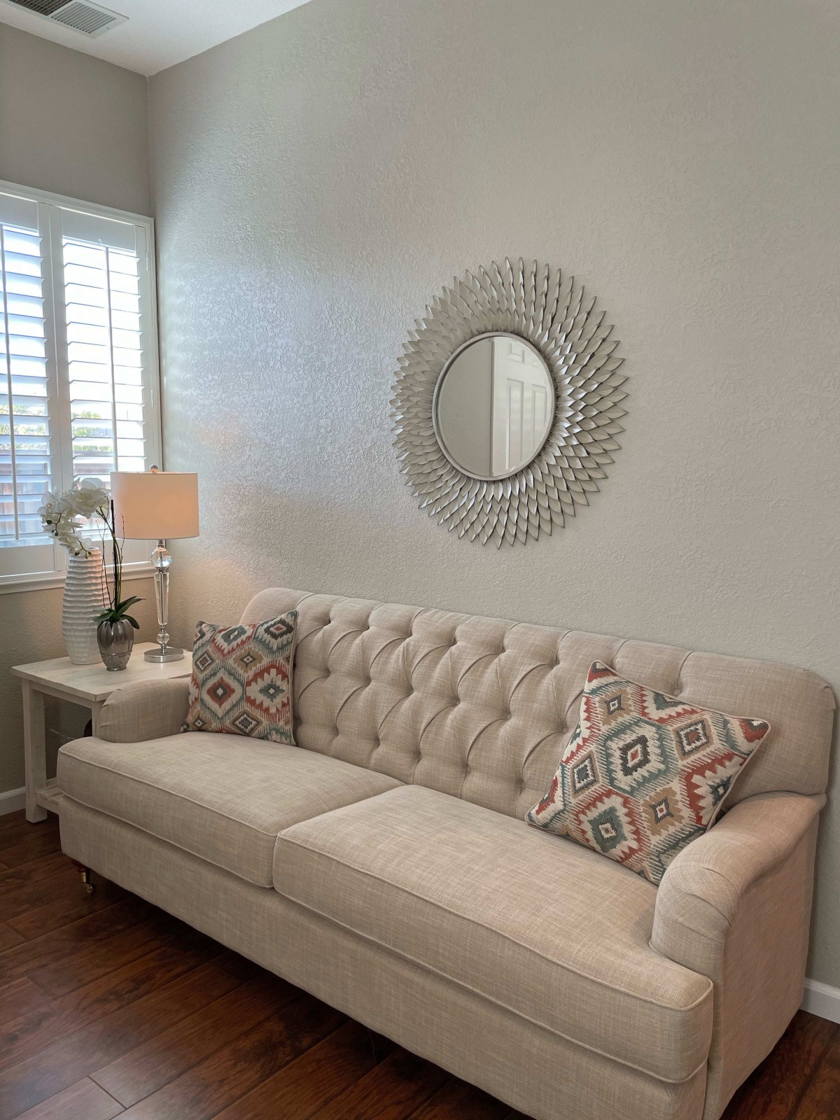 A cozy living room with a beige tufted sofa, decorative pillows, a round wall mirror, and a side table lamp by the window.