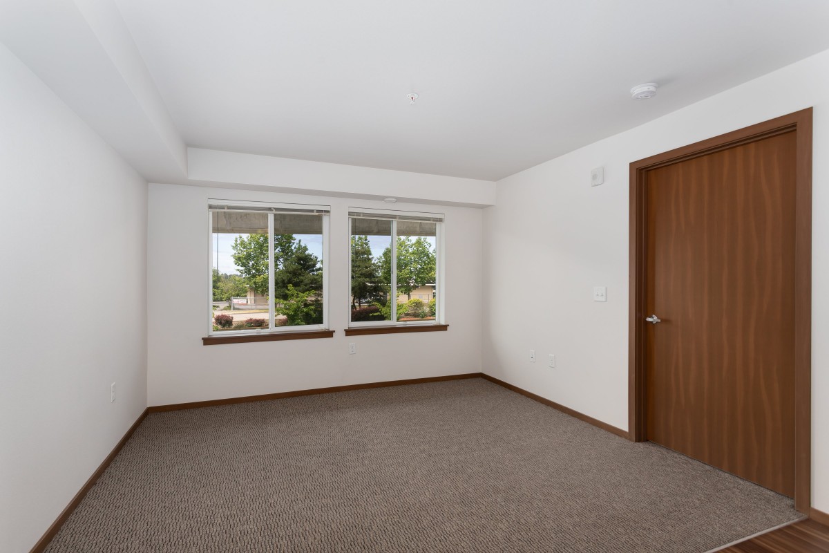 An empty room with white walls, large windows, a wooden door, and carpeted floor.