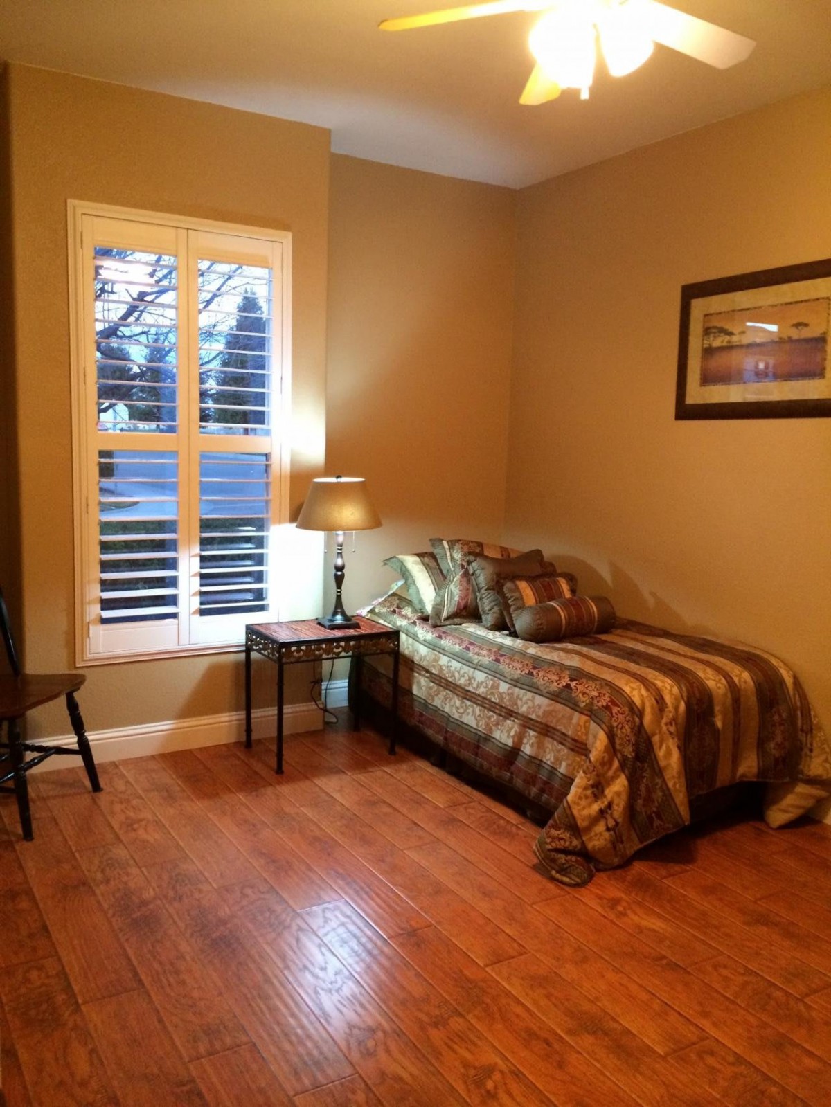 A cozy bedroom with a single bed, nightstand, and lamp, featuring warm lighting and a window with shutters.