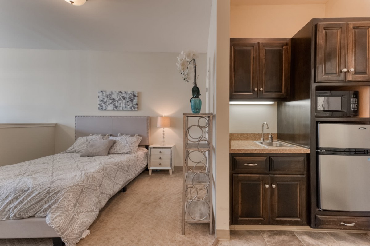 A cozy bedroom with a small kitchenette, featuring a bed, nightstand, and lamp under soft lighting.