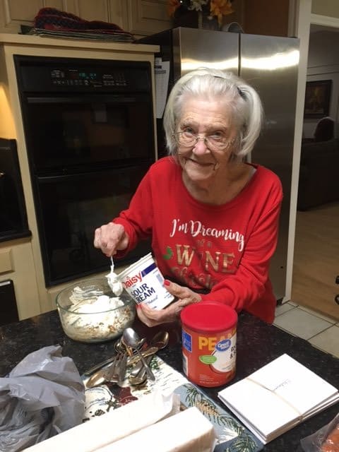An elderly woman in a red sweatshirt is joyfully preparing food in the kitchen.