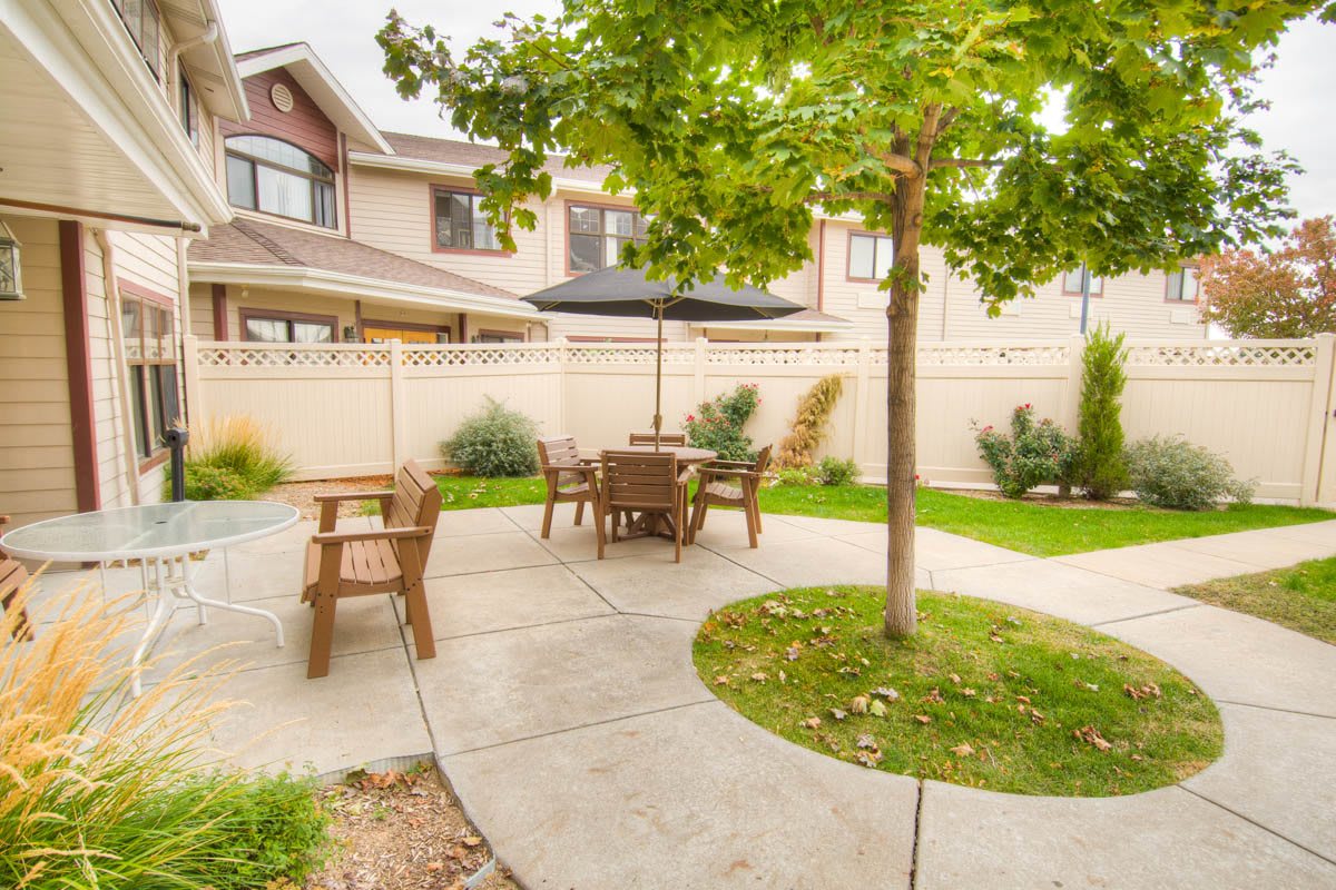 A cozy backyard patio with outdoor seating and a black umbrella, surrounded by greenery.