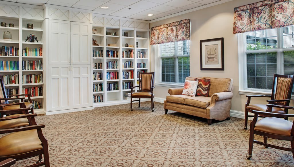 A cozy library space with a bookshelf, chair, and sofa, featuring a patterned carpet and window.
