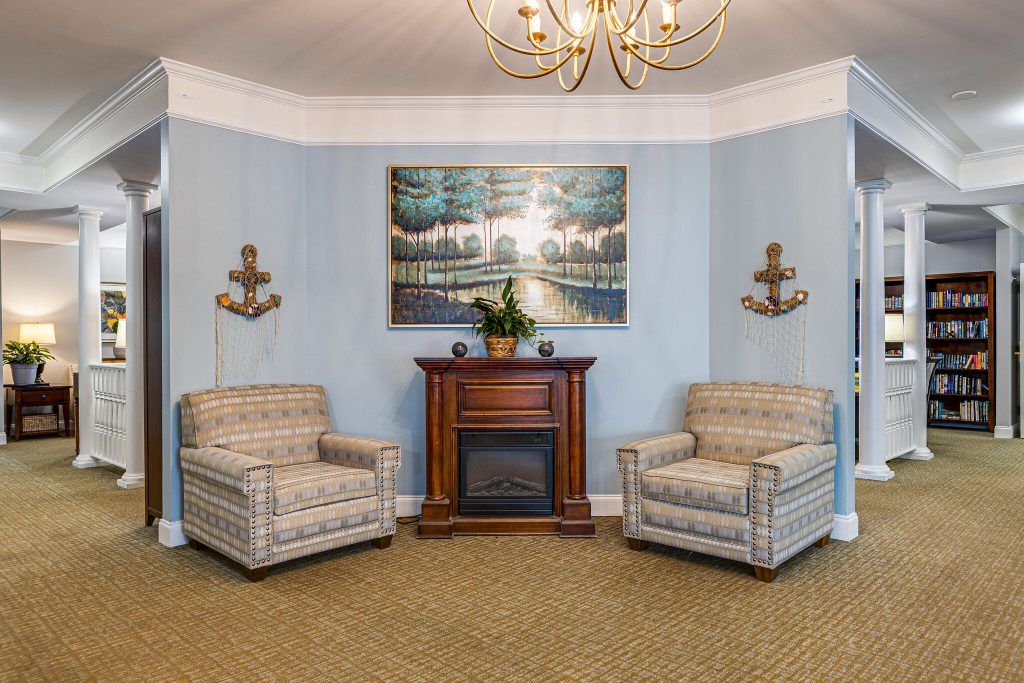 A cozy living room with two armchairs flanking a classic wooden fireplace, topped with a landscape painting and illuminated by an elegant chandelier.