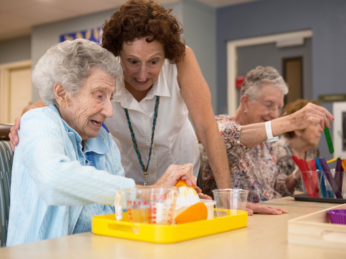 An elderly woman participates in a group activity with the help of a caregiver at a community center.