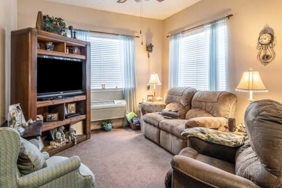 A cozy living room with a comfortable sofa, a television on a wooden bookshelf, and natural light streaming through the windows.