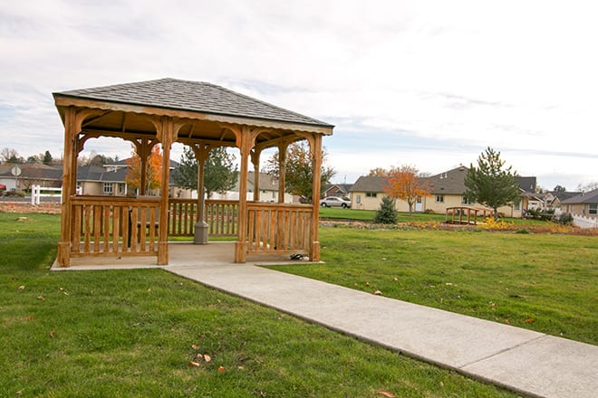 A wooden gazebo in a park with a grassy landscape and a concrete pathway.