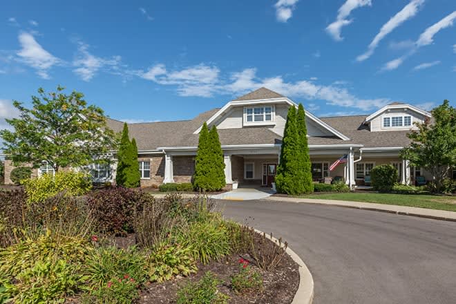 A modern residential building surrounded by landscaped gardens and a paved driveway on a sunny day.