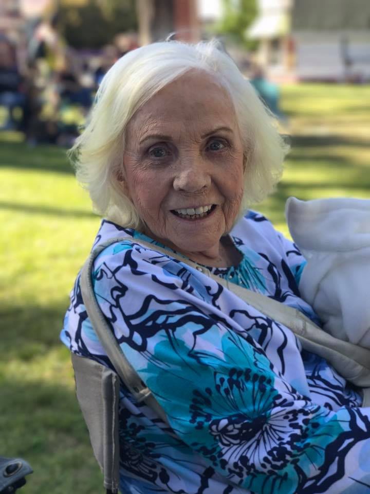 An elderly woman smiling while sitting outdoors on a sunny day.