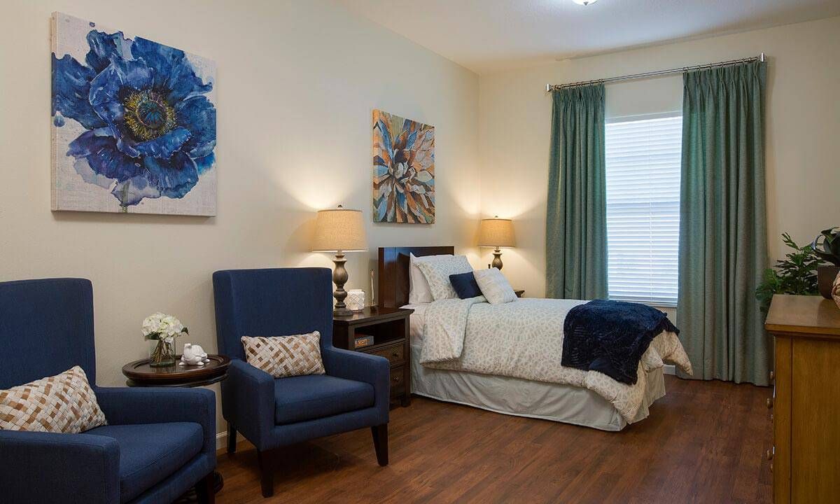A cozy and elegantly decorated bedroom featuring a bed, two blue armchairs, artwork on the walls, and soft ambient lighting.
