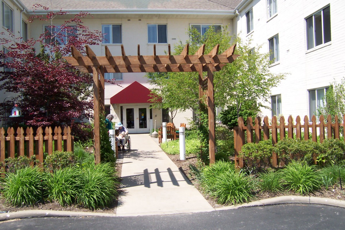 A pathway leading to a building entrance, adorned with a wooden pergola and surrounded by lush greenery.