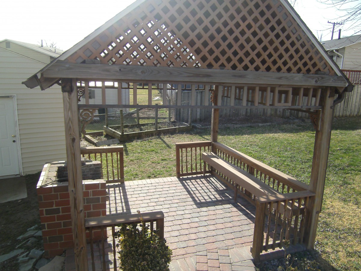 A wooden pergola with a lattice roof providing shade over a brick patio, featuring built-in benches and an open view to the backyard.