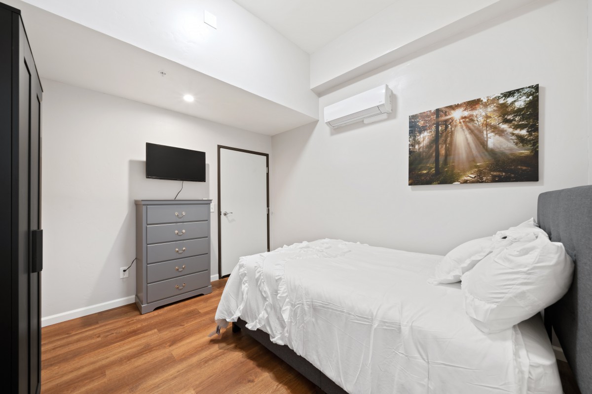 A minimalist bedroom featuring a bed, dresser, wall-mounted TV, and artwork.