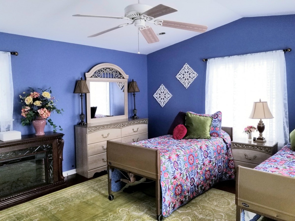 A cozy bedroom with blue walls, featuring a single bed adorned with floral bedding and decorative pillows. The room includes a dresser with a mirror, a ceiling fan, and various decorative elements.