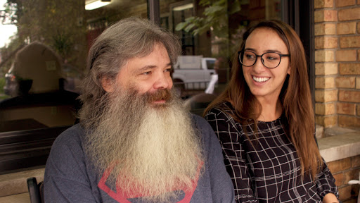A man with a long beard and a woman wearing glasses share a joyful moment outdoors.