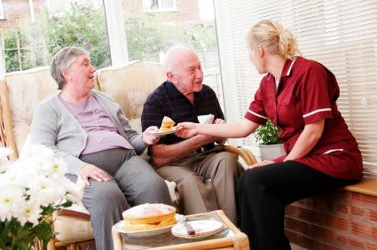 A caregiver is assisting two elderly individuals in a comfortable indoor setting.