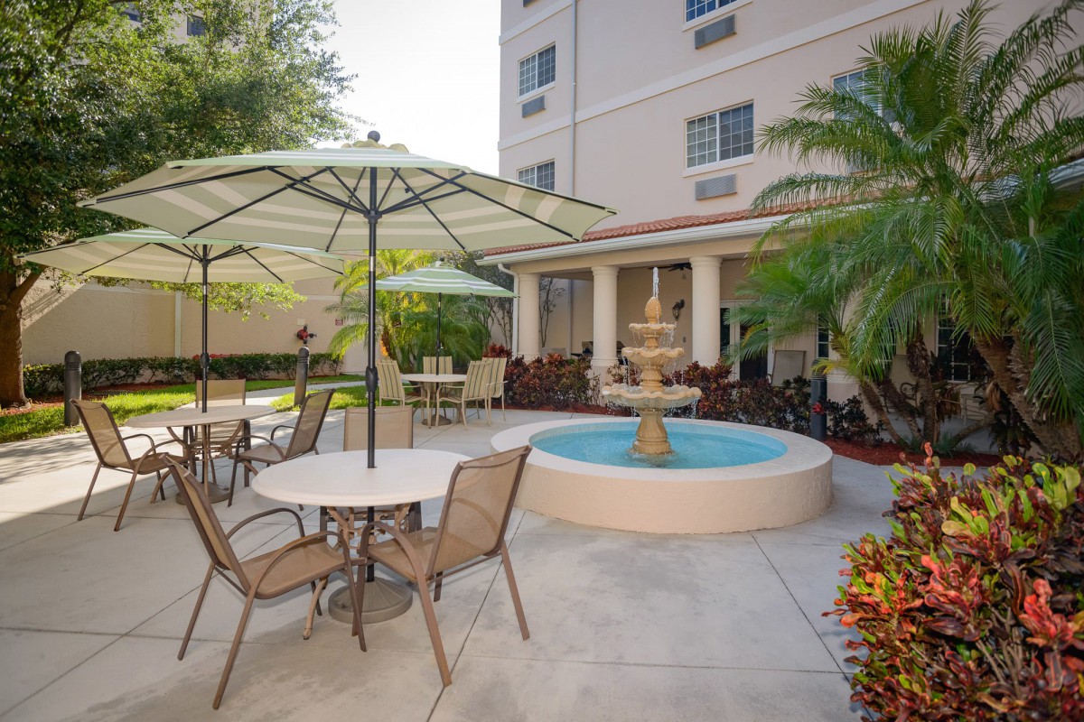 A cozy outdoor patio area with tables, chairs, and umbrellas surrounding a central fountain, adjacent to a building with palm trees providing shade.