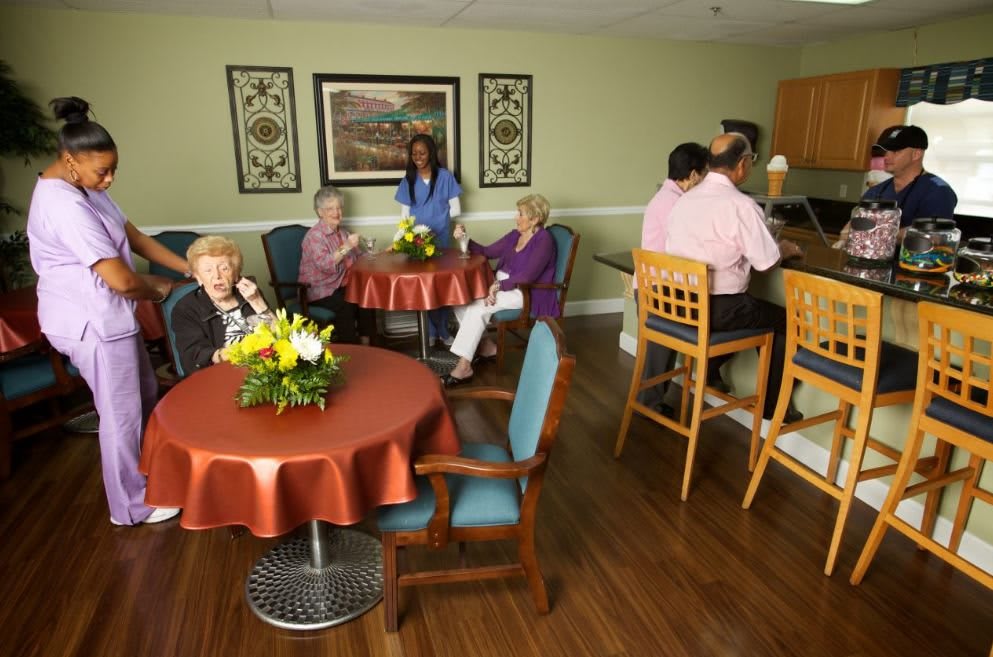 Residents and staff socializing in a care center common area with tables and flowers.