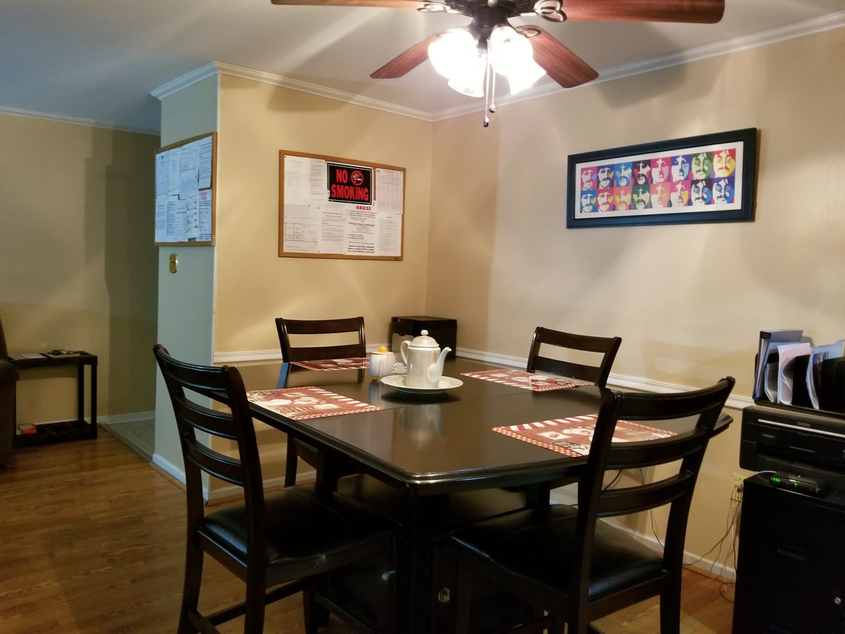 A cozy dining room featuring a dark wooden table with four chairs, adorned with placemats and a centerpiece, under a ceiling fan and surrounded by artwork on the walls.