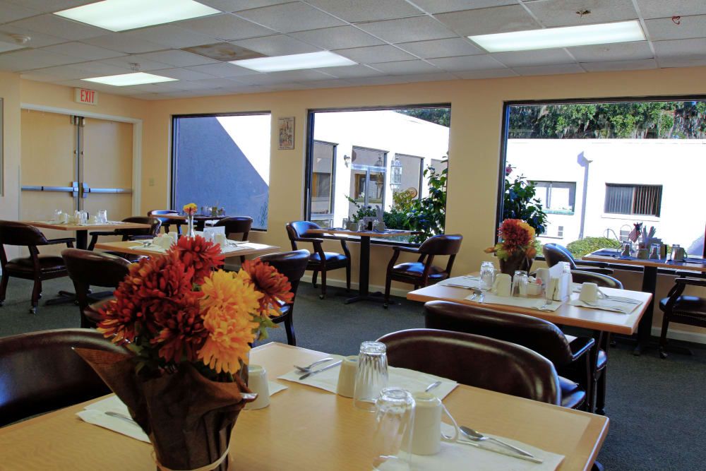 A bright dining area with tables set for a meal, featuring floral decorations and large windows.
