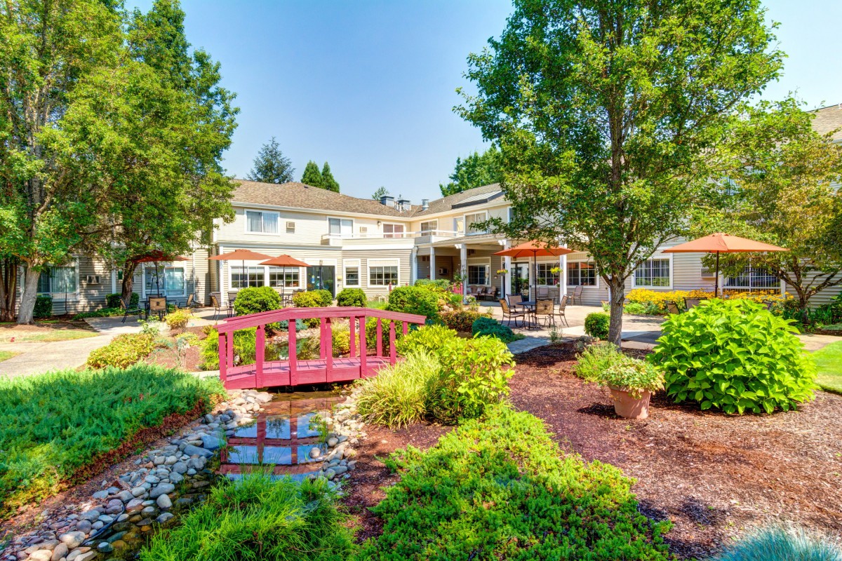 A beautiful garden with a small bridge and lush greenery leading to a building on a sunny day.