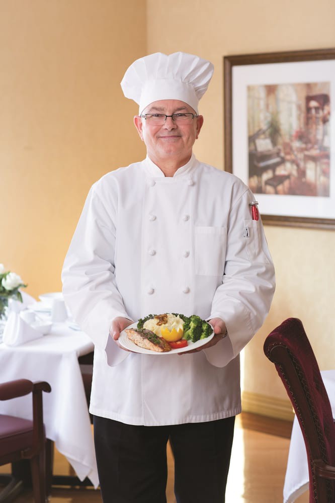 A chef in a white uniform presenting a plated dish at a restaurant.