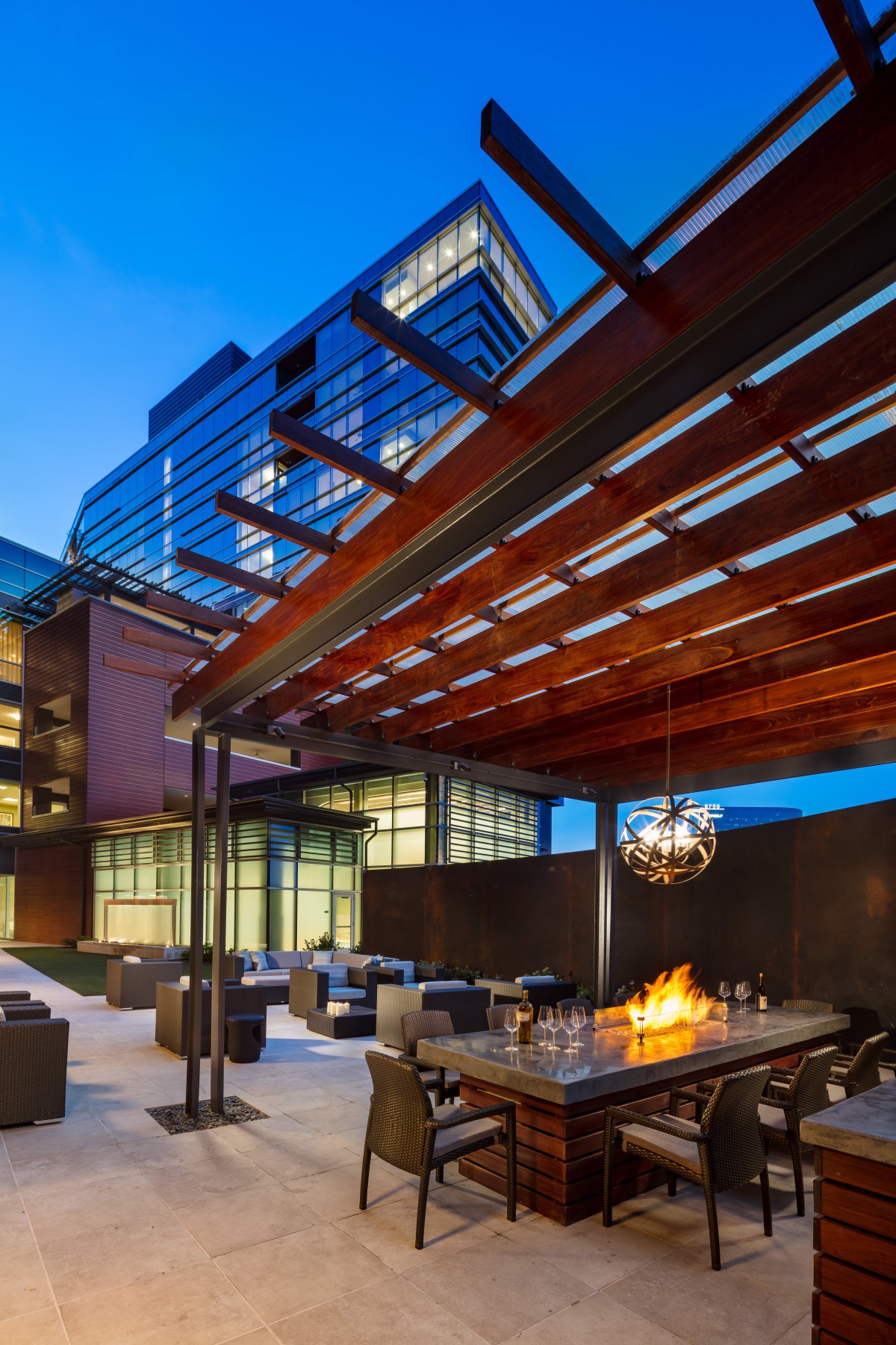 A modern outdoor patio area with a fire pit and comfortable seating, set against the backdrop of a contemporary building at dusk.