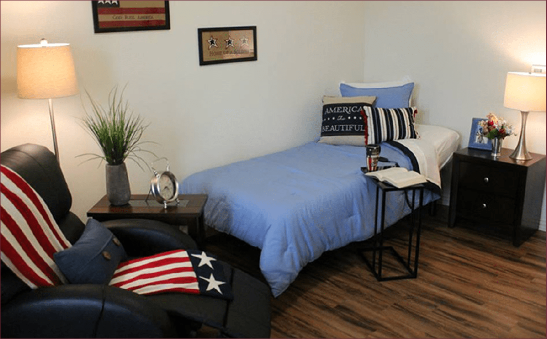 A cozy bedroom with a single bed dressed in blue bedding and decorated with patriotic-themed pillows. The room features wooden flooring, framed pictures, and a small side table with a plant.