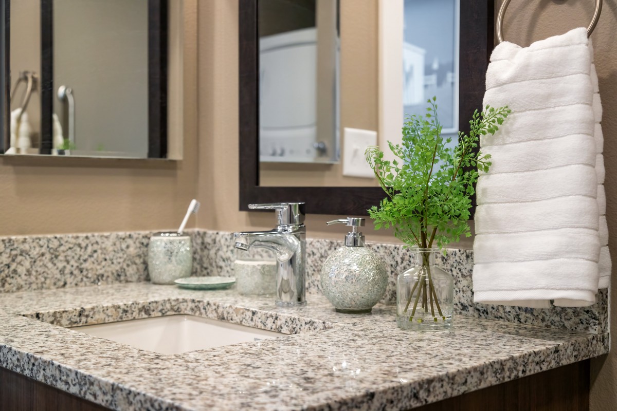 A tidy bathroom countertop featuring a granite surface, decorative jars, fresh greenery, and neatly stacked towels beside a mirror.