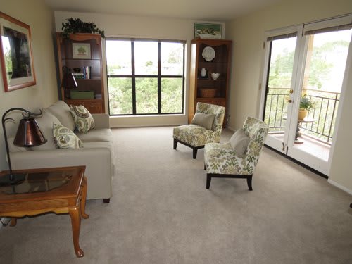 A cozy living room featuring a sofa, two chairs, and bookshelves, with a large window and sliding door leading to a balcony.