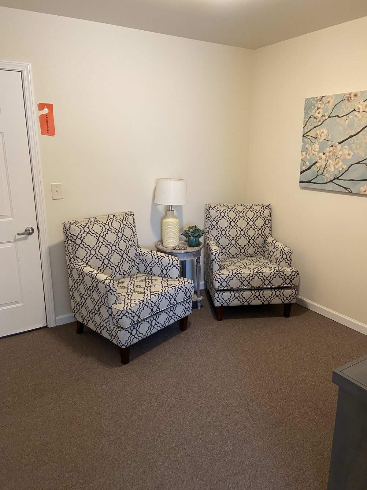 A cozy living room corner featuring two patterned chairs, a small side table with a lamp, and a floral artwork on the wall.